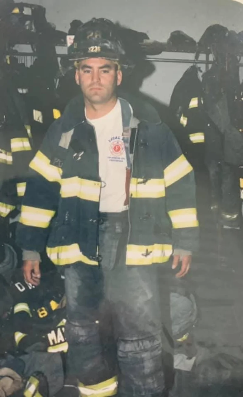 An archival photograph of a firefighter standing beside turnout gear.