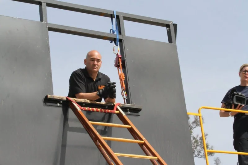 An instructor at an elevated window training prop with a ground ladder and rope equipment.