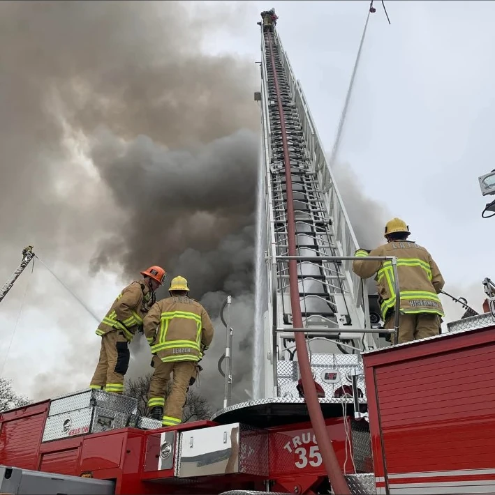 Firefighters on a red aerial ladder truck at a smoke-filled incident.
